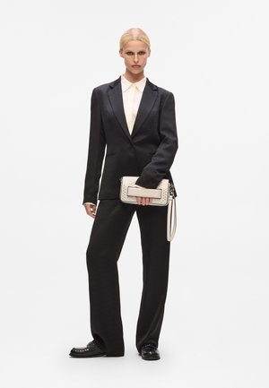 Costum negru croit, realizat dintr-un material texturat, asociat cu o cămașă crem cu guler. Ținând o geantă de mână de culoare deschisă, cu detalii de accent.
