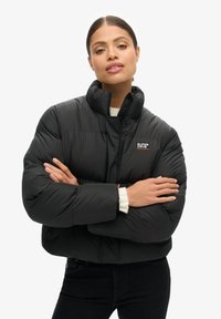 Zwarte gewatteerde jas met een hoge kraag, voorzien van een logopatch, brede mouwen en een gewatteerde textuur. Gekozen over een lichte trui.