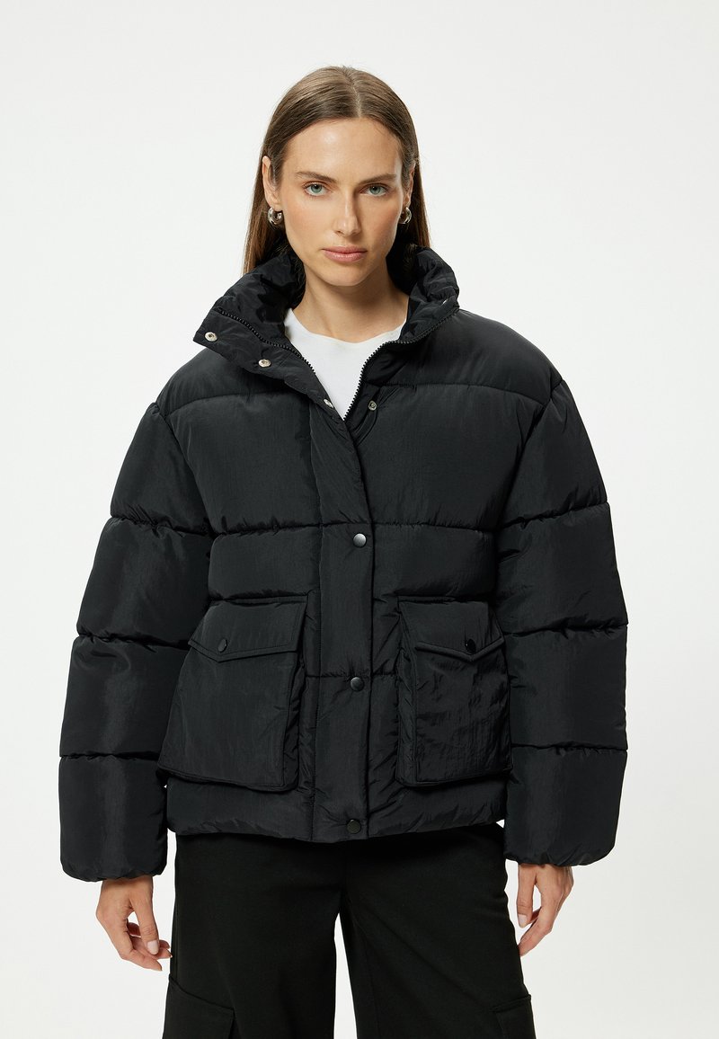 Chaqueta acolchada negra con cuello alto, botones a presión y dos bolsillos delanteros. Confeccionada con un material ligero y texturizado con un diseño acolchado.