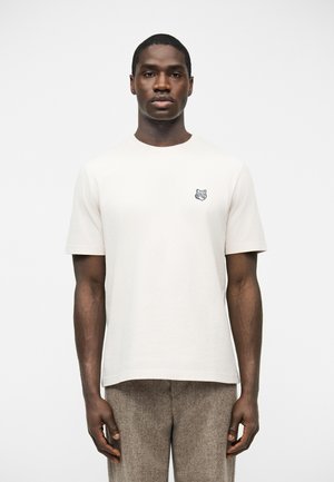 Homme debout devant un fond uni, portant un t-shirt blanc à manches courtes avec un petit logo de loup sur le côté gauche de la poitrine et un pantalon marron.