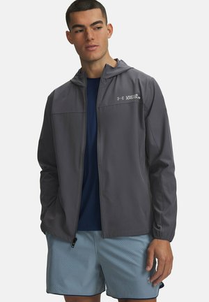 Man draagt een grijze Under Armour hoodie over een marineblauw shirt en lichtblauwe sportshorts tegen een effen achtergrond.