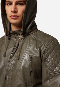 Olivgrön läderjacka med kviltad axeldesign, dragkedjeförsedd bröstficka och justerbar dragsko i huvan. Slät textur.