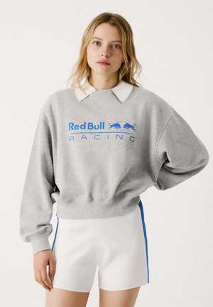 Sweat-shirt gris avec impression bleue "Red Bull Racing", col blanc et coupe oversize. Associé à un short blanc avec des rayures latérales bleues.