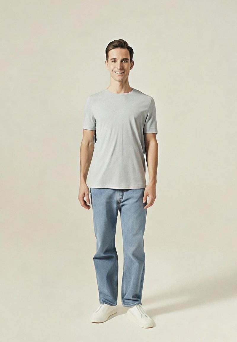 Hombre sonriendo de pie, vistiendo una camiseta gris clara, jeans azul claro y zapatillas deportivas blancas frente a un fondo beige liso.