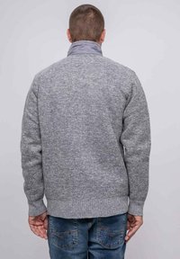 Homme de dos portant un pull en tricot gris à col montant et un jean en denim bleu, sur fond clair.