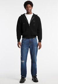 Sudadera con cremallera negra de textura con capucha, combinada con jeans azules desgastados y zapatos negros, ofreciendo un look casual en capas.
