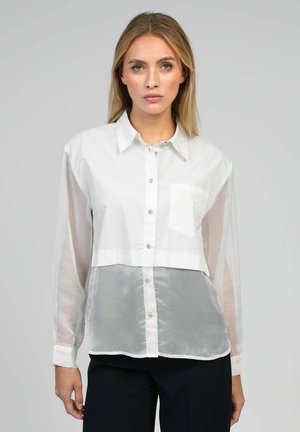 Femme blonde portant un chemisier blanc transparent à manches longues avec une poche poitrine et un pantalon noir, debout devant un fond uni.