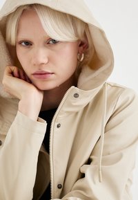 Veste imperméable beige avec une capuche texturée doublée en polaire, des boutons-pression argentés et un cordon de serrage ajustable. Vue en gros plan.