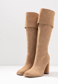 Bottes hautes jusqu'au genou en daim beige avec un revers, un bout carré et un talon haut épais. Fermeture zippée sur le côté. Texture lisse sur toute la surface.