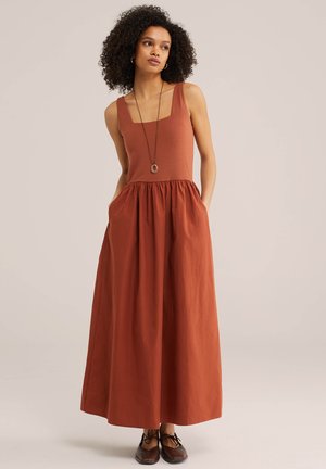 Femeie cu păr creț, purtând o rochie maxi fără mâneci, de culoare ruginiu, pantofi negri și un colier lung cu pandantiv, stând cu mâinile în buzunare.