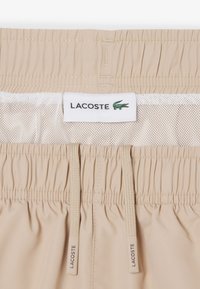 Shorts à taille élastique beige avec doublure en mesh, cordons de serrage portant l'inscription "Lacoste" et une étiquette blanche affichant le texte Lacoste et le logo crocodile vert.