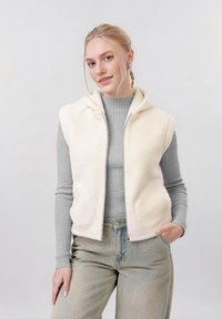 Jeune femme aux cheveux blonds tressés portant un gilet sherpa crème sur un pull côtelé gris et un jean clair, posant avec la main dans la poche.