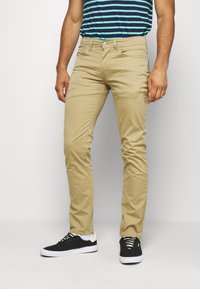 Khaki slim-fit byxor med en len textur och standard femfickorsdesign, matchade med svarta sneakers och en blårandig skjorta.