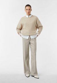 Beżowy gruby sweter z dzianiny nałożony na białą koszulę z kołnierzykiem, zestawiony z jasnoszarymi spodniami w prążki i białymi butami w szpic.