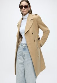 Kahekordse nööpimiskohaga kaamelivärvi mantel kuldse tooniga nuppudega, kohandatud lõikega, sile villalaadne tekstuur, kantud valge kaelusega kampsuni ja heledate teksade kohal.