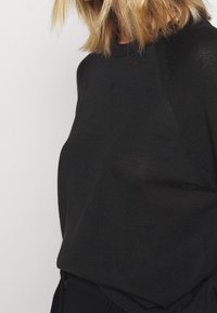 Svart stickad tröja med ett texturerat mönster och rundad halsringning, i en avslappnad passform, uppställd mot en neutral bakgrund.