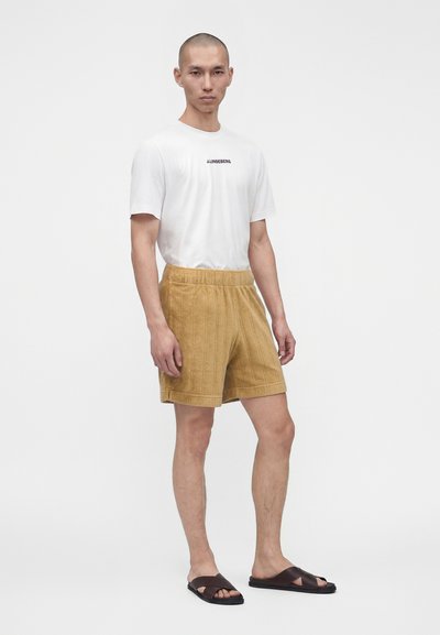 Homme debout portant un t-shirt blanc, un short texturé couleur moutarde, et des sandales marron foncé à brides croisées sur fond blanc uni.