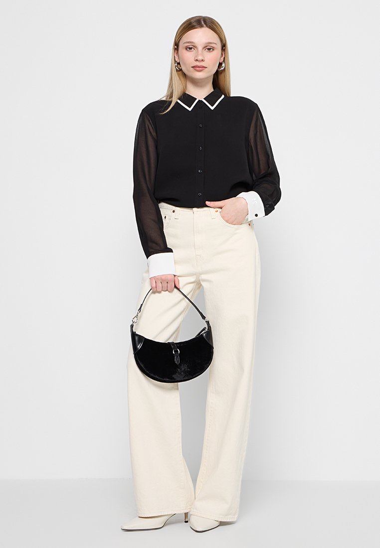 Schwarze transparente Bluse mit weißem Kontrastkragen, hochtaillierte cremefarbene weit geschnittene Jeans, hält eine schwarze, geschwungene Handtasche und trägt spitz zulaufende weiße High Heels.