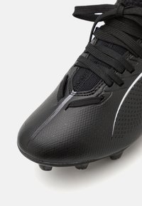 Bota de fútbol negra con material texturizado, presenta un diseño de cordones, costuras prominentes y una franja blanca en el costado.