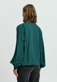 Blusa verde con texture e maniche lunghe a sbuffo, con strisce verticali e polsini con bottoni, indossata sopra jeans scuri.