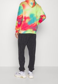 Mikina s kapucí s tie-dye vzorem v živých barvách (růžová, zelená, modrá) spárovaná s tmavými, mírně obnošenými joggingovými kalhotami a teniskami.