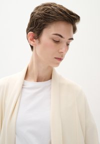 Krämfärgad, lätt cardigan med öppen front och långa ärmar, bärs över en enkel vit t-shirt. Slät textur och minimalistisk design.