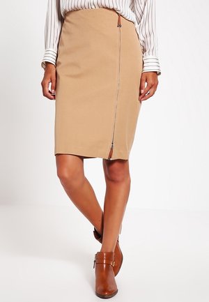 Beige knælængde nederdel med front lynlåsdetaile, båret med brune ankelstøvler og en hvid bluse med sorte lodrette striber.