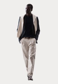 Leichtgewichtiger Blazer in Marineblau, locker drapiertem cremefarbenem Schal, schwarzem Hemd, beigefarbenen, plissierten Hosen und braunen Schuhen schaffen einen modernen, geschichteten Look.