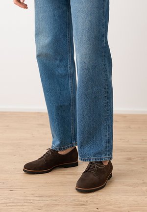 Person wearing blue jeans and dark brown suede lace-up shoes standing on a light wooden floor.