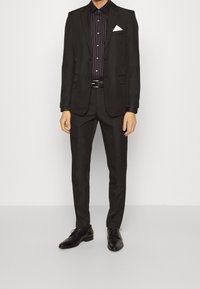 Completo nero con giacca e pantaloni su misura, abbinato a una camicia a righe bianco e nere. Da notare il fazzoletto bianco nel taschino e le scarpe nere.