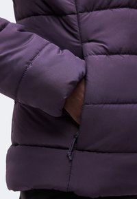 Main partiellement dans la poche d'une doudoune matelassée violet foncé avec des coutures visibles et un tire-zip.