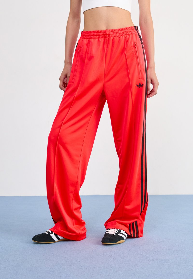 Červené športové nohavice vyrobené z lesklého materiálu. Majú voľný strih, elastický pás, bočné vrecká a čierne pruhy adidas po stranách.