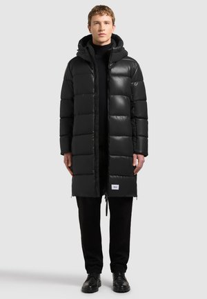 Joven con un abrigo negro acolchado largo con capucha, pantalones negros y zapatos negros, de pie frente a un fondo liso gris claro.