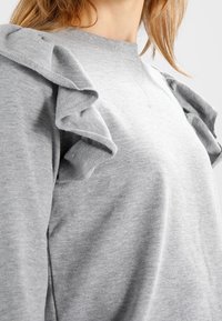 Harmaa collegepaita, jossa on olkapäillä röyhelökoristeita. Valmistettu pehmeästä kankaasta, pyöreällä kaula-aukollla ja sileällä tekstuurilla.