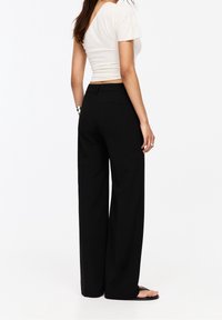 Pantaloni neri a gamba larga con una texture liscia, dotati di tasche posteriori e una vita aderente, abbinati a una maglietta aderente color crema.