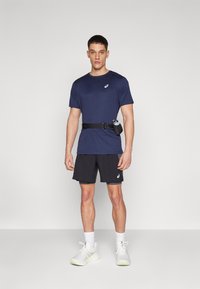 Marineblaues Sportshirt, schwarze Shorts und weiße Laufschuhe. Ein schwarzer Gürtel mit einem Flaschenhalter ist um die Taille getragen.