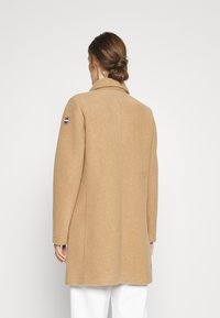 Wollmantel in Kamelbraun mit hohem Kragen, langen Ärmeln und leicht taillierter Silhouette. Verfügt über ein Logo-Patch auf dem linken Ärmel.