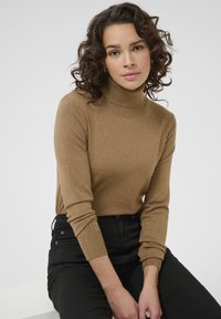 Brązowy sweter z kołnierzem typu golf, z długimi rękawami, wykonany z miękkiego dzianinowego materiału, w zestawieniu z czarnymi spodniami, na neutralnym tle.