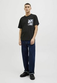 Camiseta de algodón negra con un diseño de bolsillo gráfico blanco, combinada con jeans azules y zapatos negros, con un corte relajado y mangas cortas.