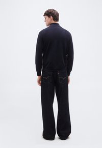 Άνδρας που βλέπει προς τα πίσω, φοράει μαύρη μπλούζα με μακριά μανίκια και άνετο σκουρομπλε τζιν με ραμμένες πίσω τσέπες, σε απλό λευκό φόντο.