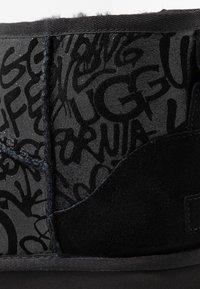 Botín negro con textura de ante y brillo, que presenta atrevidos diseños en estilo grafiti en negro. Incluye un suave forro de piel negra y detalles cosidos.