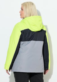 Kobieta z blond włosami zwrócona bokiem, ubrana w zimową kurtkę z kapturem w neonowo-żółtym, czarnym i szarym kolorze, stojąca na tle białej ściany.