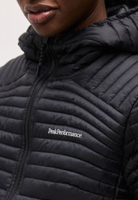 Close-up van een persoon die een zwart doorgestikt jack draagt met een ritssluiting en het logo van "Peak Performance" op de borst.