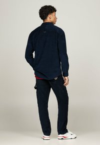 Chemise en velours côtelé bleu marine à manches longues avec un détail de boucle au dos, assortie à un pantalon cargo et des baskets blanches avec des accents rouges.
