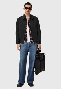 Afslappet sort jakke med elastiske manchetter over en blomsterprintet skjorte, parret med løse blå jeans og en sort tote bag. Fremhæver mørke solbriller.