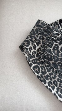 Tissu en denim imprimé léopard plié, présentant des motifs noirs et gris. Détails de surpiqûre sur les bords. Fond en tissu léger et texturé.