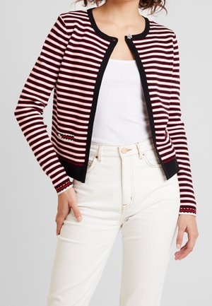 Vrouw draagt een rood, zwart en wit gestreepte cardigan over een witte tanktop met een hoge taille witte spijkerbroek tegen een effen achtergrond.