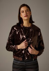 Brown sequined bomber jacket with a ribbed collar and cuffs, zip closure, and a slightly cropped fit, worn over a patterned top.