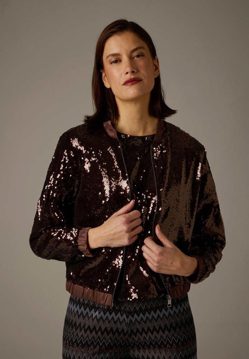 Brown sequined bomber jacket with a ribbed collar and cuffs, zip closure, and a slightly cropped fit, worn over a patterned top.