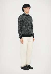 Maglione nero a motivo con collo alto, caratterizzato da un design con logo ripetuto. Indossato con pantaloni color crema e scarpe nere.